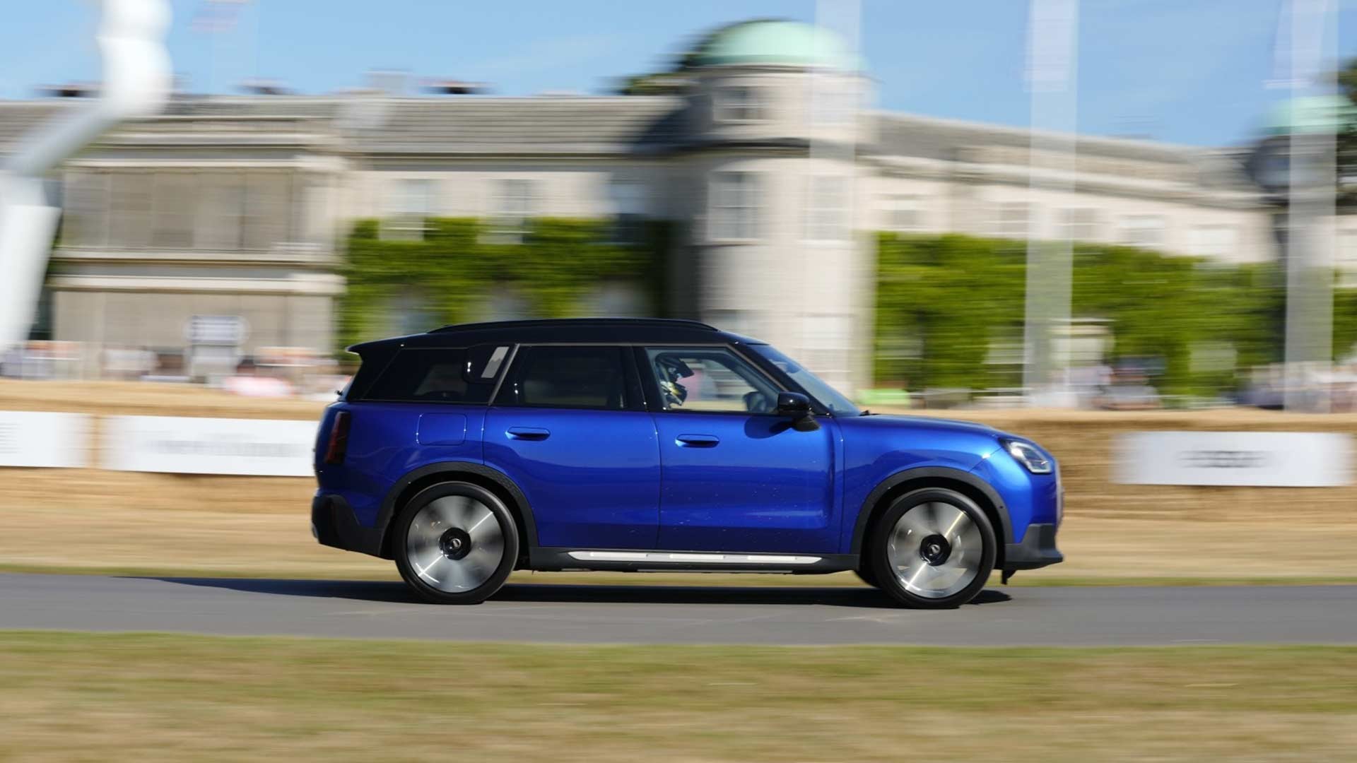  A blue SUV speeds past a historic building under a clear sky, evoking excitement. A modern sculpture and flags are visible in the blurred background.